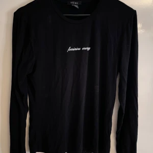 Svart långärmad tröja från Amisu - Svart långärmad tröja från Amisu med vit broderad text 'feminine energy' på bröstet. Tröjan har en klassisk rund halsringning och är gjord i mjuk bomull, perfekt för en clean och stilren look.