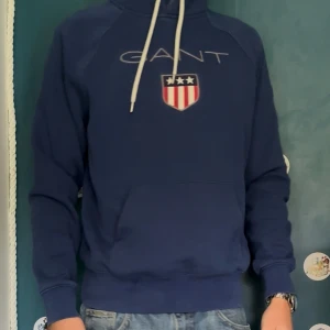 Blå hoodie från GANT med tryck - Säljer en marinblå hoodie från GANT med vit snörning i huvan och ett stort broderat GANT-tryck samt sköld med stjärnor och ränder på bröstet. Tröjan har känguruficka och ribbade muddar. Perfekt för dig som gillar klassisk och sportig stil.