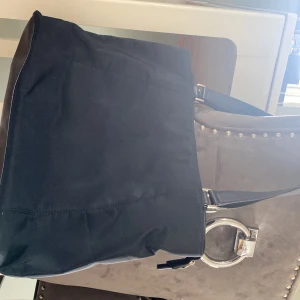 Svart axelväska med metallring - Stilren svart axelväska från WERA i polyester med snygg metallring och låsdetalj framtill. Väskan har dragkedja upptill och flera innerfack, perfekt för att hålla ordning på dina grejer. Enkel och modern design som passar till många outfits.