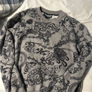 Grå mönstrad sweatshirt Versace Jeans - Säljer en grå sweatshirt från Versace Jeans Couture med svart barockinspirerat mönster över hela tröjan. Tröjan har rund halsringning, ribbade muddar och är långärmad. Perfekt statement-piece för dig som gillar lyxig streetstyle.
