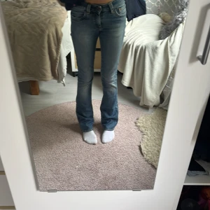 Blå bootcut jeans från Crocker W24 L32 - Snygga blå jeans från Crocker, modell CR-334, med bootcut passform och klassiska fickor bak med gul söm. Jeansen har låg midja och är gjorda i mjukt denim med slitningar för en cool look. Perfekta för dig som gillar en tidlös stil med lite edge.