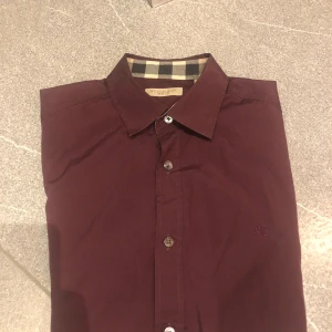 Vinröd skjorta från Burberry Brit XS - Säljer en vinröd skjorta från Burberry Brit i storlek XS. Skjortan har klassiskt rutigt mönster på insidan av kragen och manschetterna, broderad logga på bröstet och knäppning framtill. Slim passform och långa ärmar. Materialet är mjuk bomull. Säljer då den är lite för liten. Pris kan diskuteras!