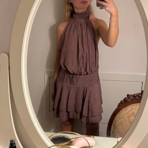 Klänning med volang - Supersnygg kortklänning med hög hals och knyt i nacken. Klänningen har volangig kjol, markerad midja och öppen rygg. Perfekt för dig som gillar en chic och trendig look. Materialet känns lätt och luftigt.