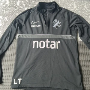 Svart AIK Nike träningströja med dragkedja - Svart långärmad träningströja från Nike med AIK:s klubbmärke, sponsorer och dragkedja vid halsen. Tillverkad i Dri-Fit material som andas. Perfekt flr träning eller en avslappnad stil. 