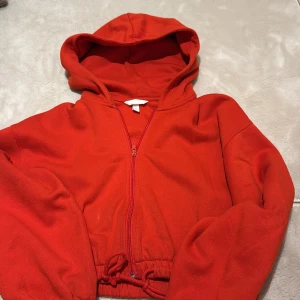 Röd hoodie med dragkedja och snörning - Röd hoodie från H&M med dragkedja framtill, huva och snörning i midjan. Tröjan är croppad och har långa ärmar med muddar. Perfekt för dig som gillar en chill och sportig stil.