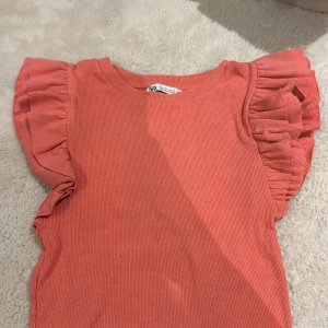 Koralldröm volangtopp från Zara - Supersöt korallfärgad topp från Zara med ribbad struktur och volangärmar. Toppen är ärmlös med breda volanger som ger en extra touch. Perfekt för dig som vill sticka ut med färg och detaljer. Materialet är mjukt och stretchigt för en bekväm passform.