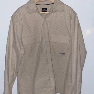 Beige overshirt från Essentials  - Säljer en beige overshirt från Essentials i storlek L. Skjortan har diskret randigt mönster, två stora bröstfickor med lock och knäppning framtill. Tillverkad i mjuk manchester som ger en chill och trendig vibe. Perfekt lager-på-lager-plagg för din streetwear-look.