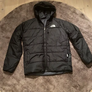 Svart pufferjacka från The North Face - Svart pufferjacka från The North Face med huva och dragkedja framtill. Jackan har quiltad design och logga på bröstet och ryggen. Perfekt för kalla dagar med sin vadderade känsla och klassiska look.