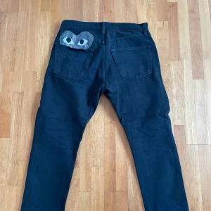 Cdg jeans - Svarta jeans med rak passform och klassisk femficksdesign. På ena bakfickan finns en unik camo-detalj med ögonmotiv som sticker ut. Jeansen har knapp- och dragkedjegylf samt bälteshällor. Perfekta för dig som vill ha något extra i din outfit.