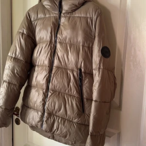 Beige pufferjacka med huva - Snygg beige pufferjacka med huva och svarta dragkedjor. Jackan har quiltad design, två fickor med dragkedja och en patch på ärmen. Perfekt för kyliga dagar och riktigt skön att bära.från zara💕