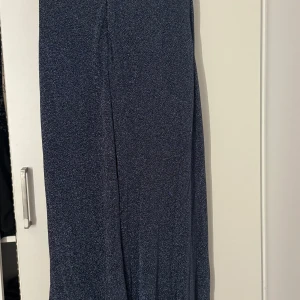 Glittrig marinblå långklänning - Snygg marinblå långklänning med glittrigt tyg och draperad detalj i midjan som pryds av små pärlor. Klänningen har en elegant och figurnära siluett, perfekt för att sticka ut. Materialet är mjukt och följsamt, vilket ger en lyxig känsla.