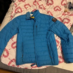 Blå dunjacka från Parajumpers - Snygg blå dunjacka från Parajumpers med klassisk patch på ärmen och gul detalj vid kragen. Jackan har dragkedja framtill, två sidofickor med dragkedja och quiltad design. Insidan är ljusgrå och jackan är lätt och smidig, perfekt för kyliga dagar.