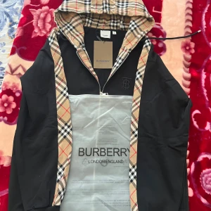 Burberry vindjacka med klassiskt rutmönster - Snygg svart vindjacka från Burberry med ikoniskt beige, svart och rött rutmönster på huva och insida. Jackan har dragkedja framtill, justerbar huva och broderad Burberry-logga på bröstet. Perfekt för dig som vill ha en stilren och tidlös look.