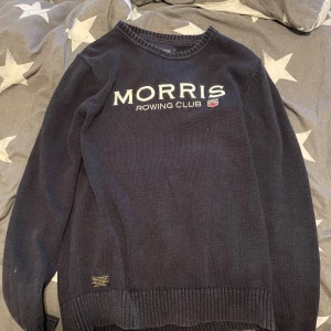Morris Rowing Club stickad tröja marinblå - Säljer en marinblå stickad tröja från Morris Rowing Club med broderad logga och liten brittisk flagga framtill. Tröjan har rund hals, ribbade muddar och är långärmad. Perfekt för dig som gillar klassisk och sportig stil.