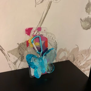 Blå fjäril nyckelring med karbinhake - Snygg nyckelring med en stor blå fjäril i transparent plast och texten 'Dotter' på ena vingen. Fästet är en metallicblå karbinhake som gör det enkelt att sätta fast på väskan eller nyckelknippan. Perfekt accessoar för dig som gillar färgglada detaljer.