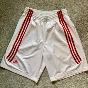 Vita Adidas BB shorts med röda ränder - Snygga vita Adidas basket shorts med klassiska röda ränder längs sidorna och Adidas-logga broderad framtill. Modellen har elastisk midja och är tillverkad i lätt polyester som andas. Perfekta för sport eller chill, med en loose passform och sportig vibe.