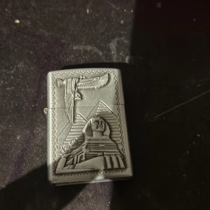 Metalltändare med Egyptiskt motiv - Stilren metalltändare med relief av Sfinxen, pyramider och en fågel. Klassisk Zippo-liknande design. Tändaren är i gott skick med mindre spår av användning, perfekt för samlare eller som present.