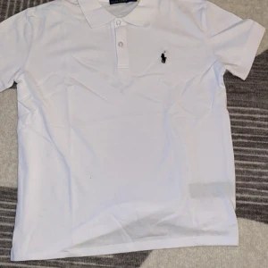 Vit pikétröja från Polo Ralph Lauren - Vit pikétröja från Polo Ralph Lauren med klassisk krage, korta ärmar och knäppning framtill. Tröjan har den ikoniska broderade loggan på bröstet och är tillverkad i 100% bomull för en skön och luftig känsla. Ska du på semester under de kommande kalla månaderna så är detta perfekt till dig om du är 175 och under pris kan diskuteras vid snabbaffär!!