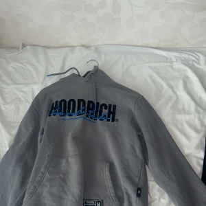 Grå Hoodrich mjukisbyxor med tryck - Snygga grå mjukisbyxor från Hoodrich med svart och blå logga på benet och patch-detalj vid fickan. Byxorna har snörning i midjan och är gjorda i mjukt bomullsmaterial. Perfekta för en chill och avslappnad stil.