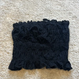 Svart bandeau tubtopp från BikBok - Säljer en svart bandeau tubtopp från BikBok i storlek XS. Toppen är smockad med volangkant upptill och nedtill, vilket ger en söt och trendig look. Perfekt att matcha med höga jeans eller kjol för en snygg outfit. Kontakta för fler bilder🩷