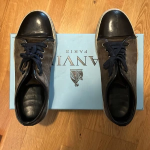 Mörkgrå sneakers från Lanvin UK8 - , Snygga sneakers från Lanvin i  mocka med svarta skinn-detaljer på tå och häl. Skorna har mörkblåa snören, . Perfekt för dig som gillar stilrena och exklusiva sneakers med en lyxig känsla. Skriv för fler bilder eller frågor! litet skrapmärke på ena captoen. 