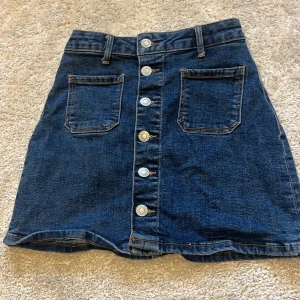 Mörkblå jeanskjol från Denim - Snygg mörkblå jeanskjol från &Denim med raka linjer, två stora fickor framtill och knappar längs hela framsidan. Kjolen har bälteshällor och klassisk denimlook, perfekt för en cool och avslappnad stil. Kontakta för fler bilder