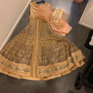 Glittrig långklänning i champagne - Superfin långklänning i champagnefärg med glittriga paljettdetaljer och broderier. Ärmlös modell med rund halsringning och vidd i kjolen. Klänningen har vackra mönster och medföljer matchande sjal i tunn tyll. Perfekt för dig som vill sticka ut med en elegant look.