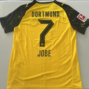 Bellingham Borussia Dortmund Puma fotbollströja - Gul och svart Borussia Dortmund fotbollströja med nummer 7 och namnet Jobe på ryggen. Tröjan har korta ärmar, Bundesliga-märke på ärmen och BVB-logga på bröstet. Tillverkad i lätt och ventilerande material, med svart grafiskt mönster på axlarna och Vodafone som sponsor. Replika, endast handtvätt gäller!