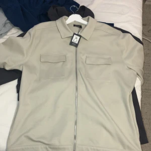 Beige overshirt med dragkedja - Stilren beige overshirt från H&M med klassisk krage och två bröstfickor med lock. Jackan har hel dragkedja framtill och är tillverkad i ett mjukt material som ger en clean look. Perfekt för lager-på-lager och enkel att matcha.