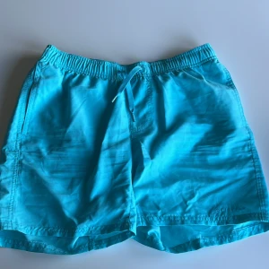 Turkosa badshorts med snörning - Snygga turkosa badshorts från Affinity med elastisk midja och snörning framtill. Shortsen har sidofickor och en bakficka med lock. Perfekta för stranden eller poolen, tillverkade i lätt och snabbtorkande syntetmaterial.