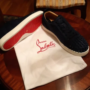 Christian Louboutin sneakers med nitar - Mörkblå sneakers från Christian Louboutin i mocka med klassisk röd sula och vita kanter. Skorna har snörning och är dekorerade med svarta nitar på tån för en edgy look. Insidan är ljusbeige och materialet känns lyxigt och mjukt.