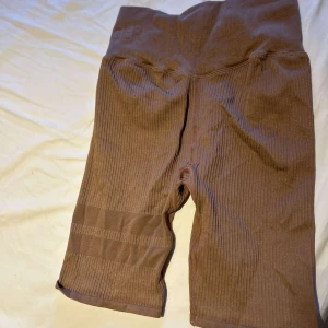 Bruna ribbade cykelshorts STRONGER - Säljer ett par bruna cykelshorts från STRONGER med hög midja och ribbad struktur. Shortsen är stretchiga och har en tight passform, perfekta för träning eller chill. Materialet är mjukt och följsamt syntet. Snygga detaljer med diskreta ränder på benen.