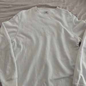 Vit sweatshirt från C.P. Company - Säljer en stilren vit sweatshirt från C.P. Company med broderad logga på bröstet. Tröjan har rund halsringning, långa ärmar och är tillverkad i mjuk bomull. Perfekt för dig som gillar clean och minimalistisk stil. Ny pris ca 1800kr
