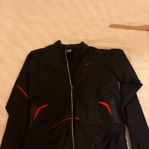 Svart vindjacka från Nike med röda detaljer - Säljer en svart vindjacka från Nike med röda detaljer och hel dragkedja framtill. Jackan har lång ärm, sportig design och är tillverkad i lätt polyester. Perfekt för dig som gillar en clean och stilren look .