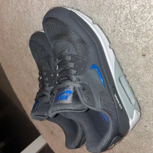 Nike Air Max 90 grå/blå sneakers - Nike Air Max 90 sneakers i mörkgrått med blå detaljer och vit sula. Ovandelen är i mesh och syntet med klassisk Air Max-dämpning. Swoosh-logga i blått på sidan och Nike Air-märke på hälen. Perfekta för dig som gillar stilrena och sportiga sneakers.