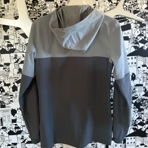 Grå vindjacka från Under Armour - Hejsan, säljer just nu en snygg tvåfärgad vindjacka från Under Armour i ljusgrått och mörkgrått. Jackan är i storlek xs och har huva, dragkedja hela vägen och en bröstficka med dragkedja. Säljer pågrund av att den var för liten på mig 