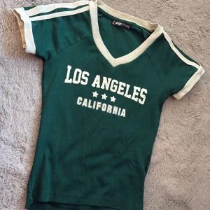 Grön Los Angeles t-shirt PB Sister XS - Cool grön t-shirt från PB Sister i storlek XS med vit text 'Los Angeles California' och tre stjärnor på bröstet. T-shirten har vita detaljer vid ärmslut, axlar och v-ringad hals. 