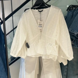 Vit omlottblus med broderade blommor - Superfin vit omlottblus med broderade blommor och v-ringning. Blusen har långa puffärmar med resår vid ärmslut och knyt i midjan. Perfekt för en fräsch och somrig look med romantiska detaljer. Använd 1 gång!