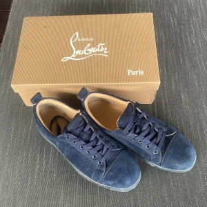 Mörkblå mocka sneakers från Louboutin - Snygga mörkblå sneakers i mocka från Christian Louboutin med klassisk röd sula. Skorna har rund tå, snörning och diskret broderad logga på hälen. Insidan är i beige läder och yttersulan är svart med röda detaljer. Perfekta för dig som gillar lyxiga och stilrena sneakers.
