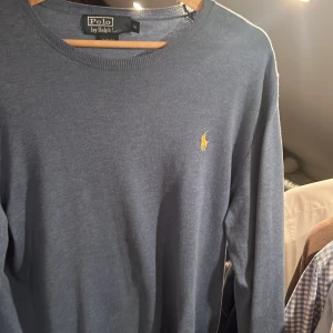 Blå tröja från Polo Ralph Lauren - Klassisk blå långärmad tröja från Polo Ralph Lauren i slim fit-modell. Tröjan har rund halsringning och den ikoniska gula broderade loggan på bröstet. Perfekt för dig som gillar stilrena och tidlösa plagg.