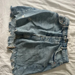 Blå jeanskjol från g perfect jeans - Säljer en blå jeanskjol från g perfect jeans i storlek 34. Kjolen är kort med råa, fransiga kanter och slitna detaljer både fram och bak. Klassisk femficksmodell med knappgylf och bälteshällor. Perfekt för en avslappnad och trendig look.