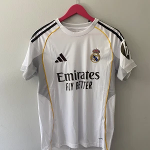 Real Madrid hemmatröja Adidas S - Säljer en officiell Real Madrid hemmatröja från Adidas i storlek S. Tröjan är vit med svarta detaljer, gula linjer och klubbmärke broderat på bröstet. Klassisk rund hals, korta ärmar och tillverkad i lätt funktionsmaterial. Perfekt för dig som älskar fotboll och vill bära klubbens färger.
