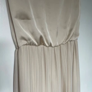 Champagnefärgad långklänning från Bik Bok - Elegant långklänning från Bik Bok i en lyxig champagnefärg. Klänningen har ett slätt, glansigt liv och ett plisserat, flowy kjolparti. Ärmlös modell med rund halsringning och markerad midja. Perfekt för dig som vill ha en stilren och sofistikerad look.🌚✨