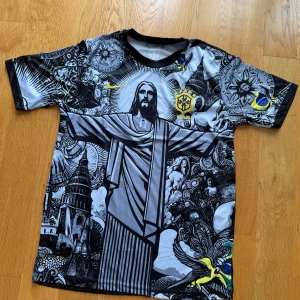 Brasil fotbollströja med Jesusmotiv Storlek M - Unik Brasilien fotbollströja från Nike med stort svartvitt tryck av Kristusstatyn och detaljerade grafiska mönster. Tröjan har korta ärmar, rund hals och inslag av gult och blått i trycket samt Brasilien-emblem på bröstet.
