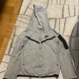 Grå Nike hoodie med dragkedja XS 122-128 - Nike hoodie i ljusgrått med hel dragkedja och svart Nike-logga på bröstet. Huva, långa ärmar och svarta detaljer längs bröstet och på ena ärmen. Praktiska fickor och sportig look, perfekt för chill eller träning.