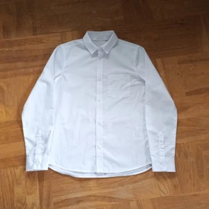 Vit skjorta från H&M, strl 152 - Klassisk vit skjorta från H&M i storlek 152. Skjortan har lång ärm, bröstficka och knäppning framtill. Tillverkad i bomul och polyester. Enkel att stryka tack vare Easy Iron-tyget. Perfekt för dig som gillar stilrena och enkla plagg.