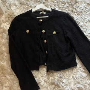 Svart kofta med guldknappar - Stilren svart kofta med rund hals och snygga guldknappar framtill. Koftan har en lätt strukturerad yta och två dekorativa fickor på bröstet. Perfekt för dig som gillar klassiska detaljer och vill ha en chic look.