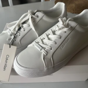 Vita sneakers från Calvin Klein - Stilrena vita sneakers från Calvin Klein med diskret logga på sidan och präglad CK-detalj runt sulan. Skorna har klassisk snörning och är tillverkade i slätt syntetmaterial för en clean look. Perfekta för dig som gillar minimalistisk och fräsch stil.