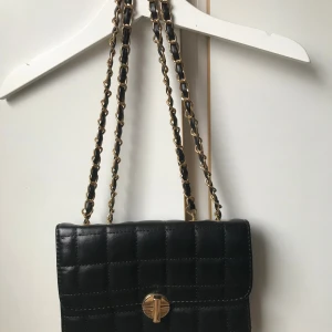 Svart quiltad axelväska med kedja - Stilren svart axelväska i quiltat skinn med guldfärgad kedjerem och snyggt vridlås framtill. Klassisk rektangulär form och elegant rutmönster. Perfekt för dig som vill ha en tidlös och trendig accessoar.