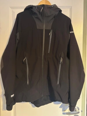 Svart och grå vindjacka från Icepeak - Säljer en svart och grå vindjacka från Icepeak med huva och flera praktiska fickor med dragkedja. Jackan har sportig look, justerbar nederkant och detaljer i grått på axlar och rygg. Perfekt för dig som gillar att vara ute och vill ha en snygg jacka med funktion. Jackan är väldigt sparsamt använd, Stl L